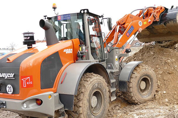 Orange-grauer Weycor-Radlader AR 660 auf einer Baustelle beim Aufnehmen von Erdmaterial mit der Schaufel. Im Hintergrund Erdhügel und Bebauung.