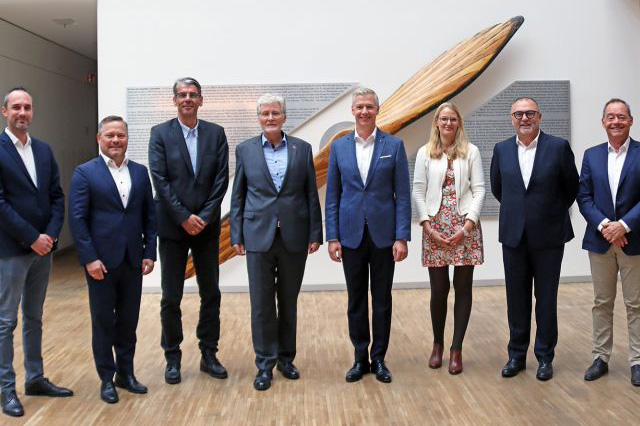Gruppenfoto mehrerer Personen in Businesskleidung vor einer Wand mit Kunstobjekt.