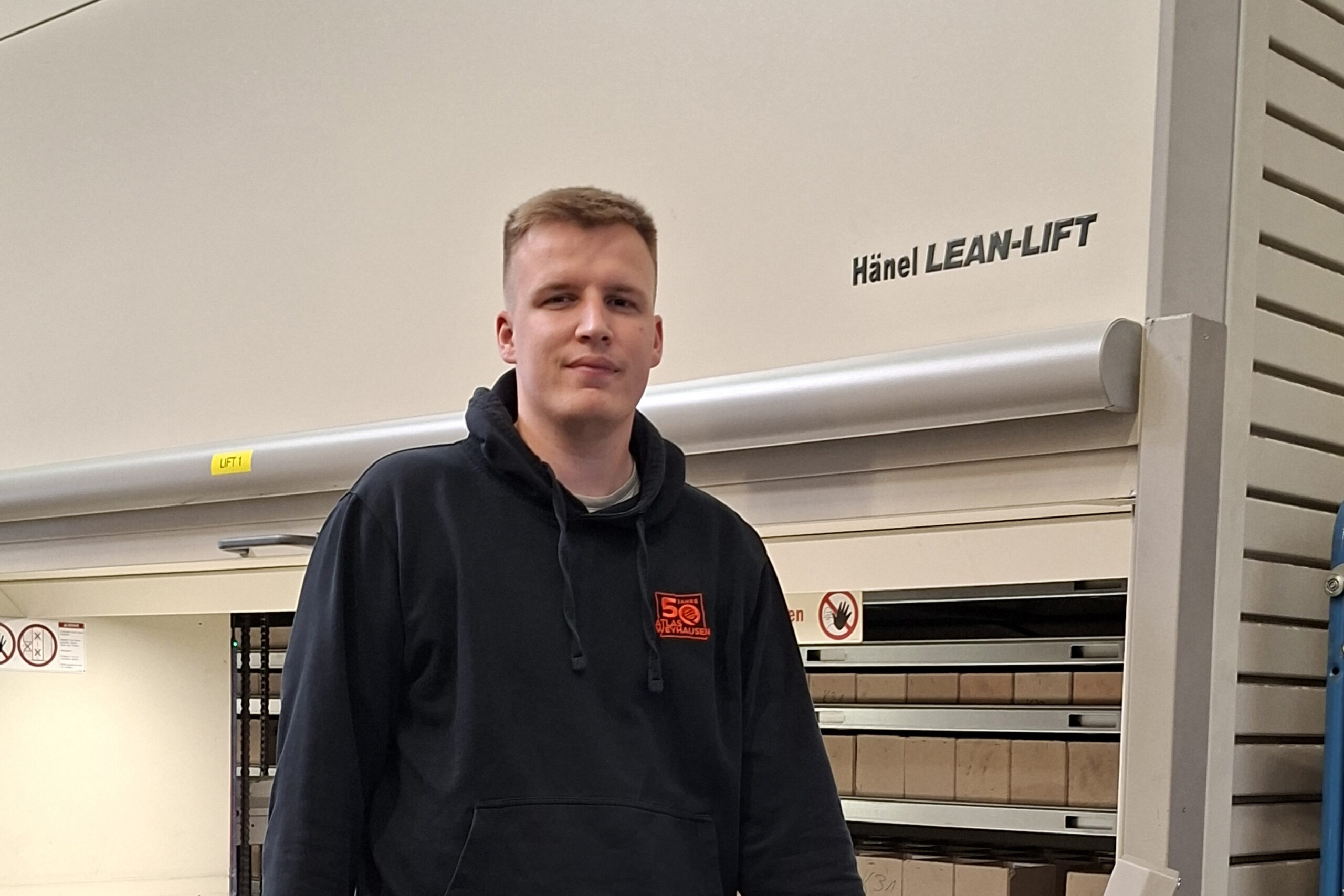Junger Mann mit kurzem Haarschnitt und dunklem Kapuzenpullover steht vor einem Hänel Lean-Lift Lagerlift. Im Hintergrund sind Fächer mit beschrifteten Lagerboxen sichtbar.