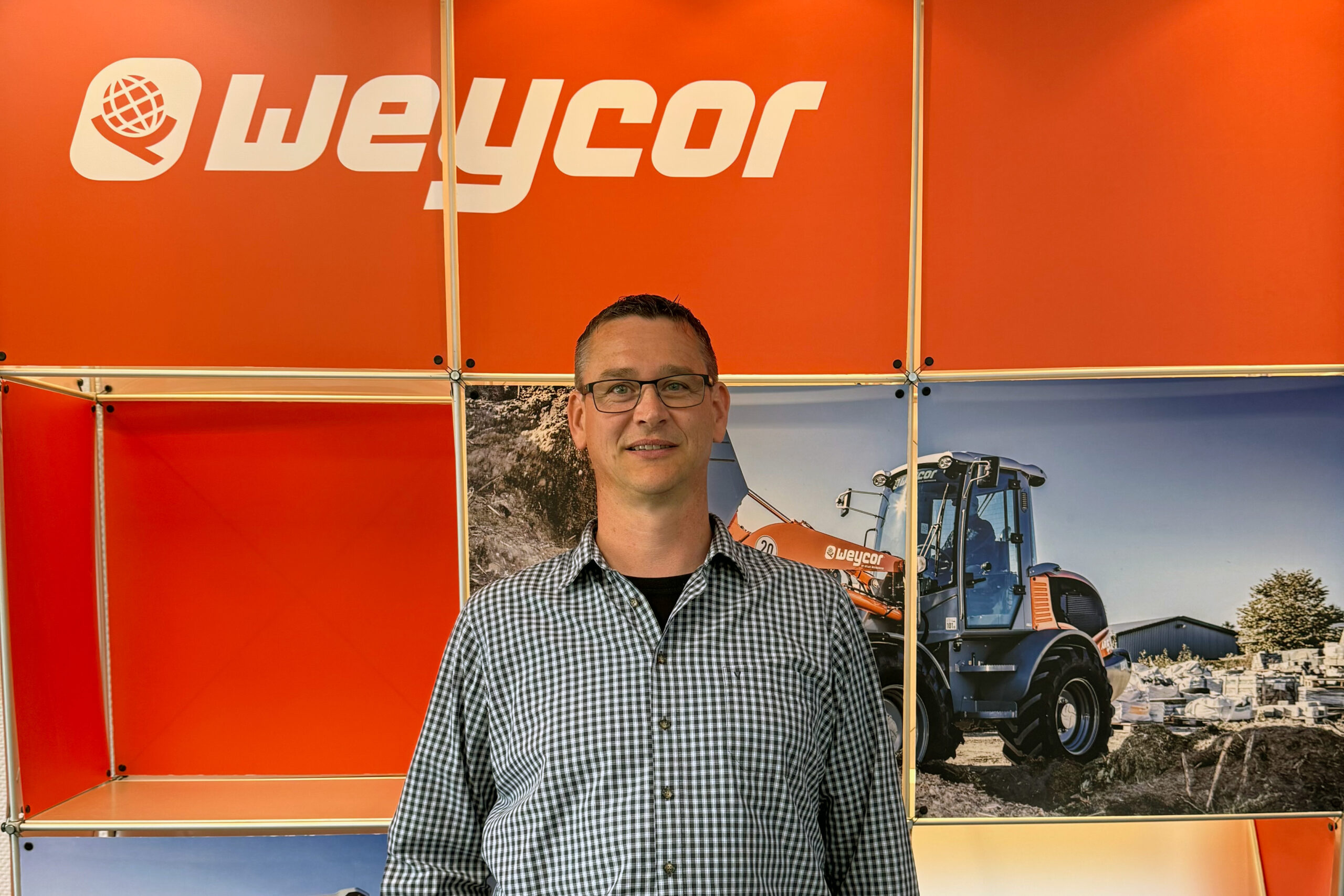 Mann mit Brille und kariertem Hemd steht vor einer orangen Messewand mit dem Weycor-Logo. Im Hintergrund ein Bild eines Weycor-Radladers auf einer Baustelle.