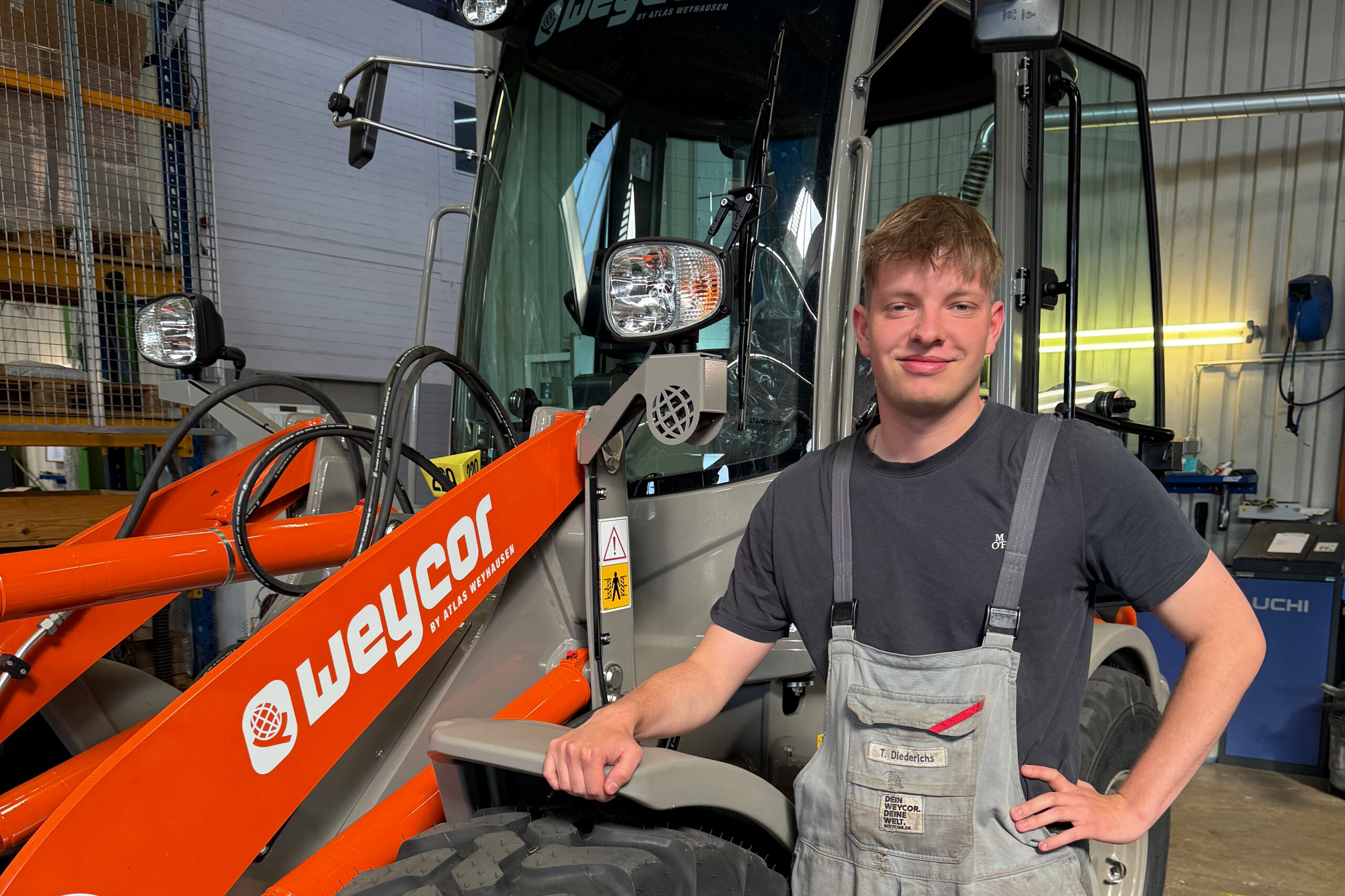 Junger Mann in Arbeitskleidung steht vor einem orange-grauen Weycor-Radlader in einer Werkstatthalle und lächelt in die Kamera.