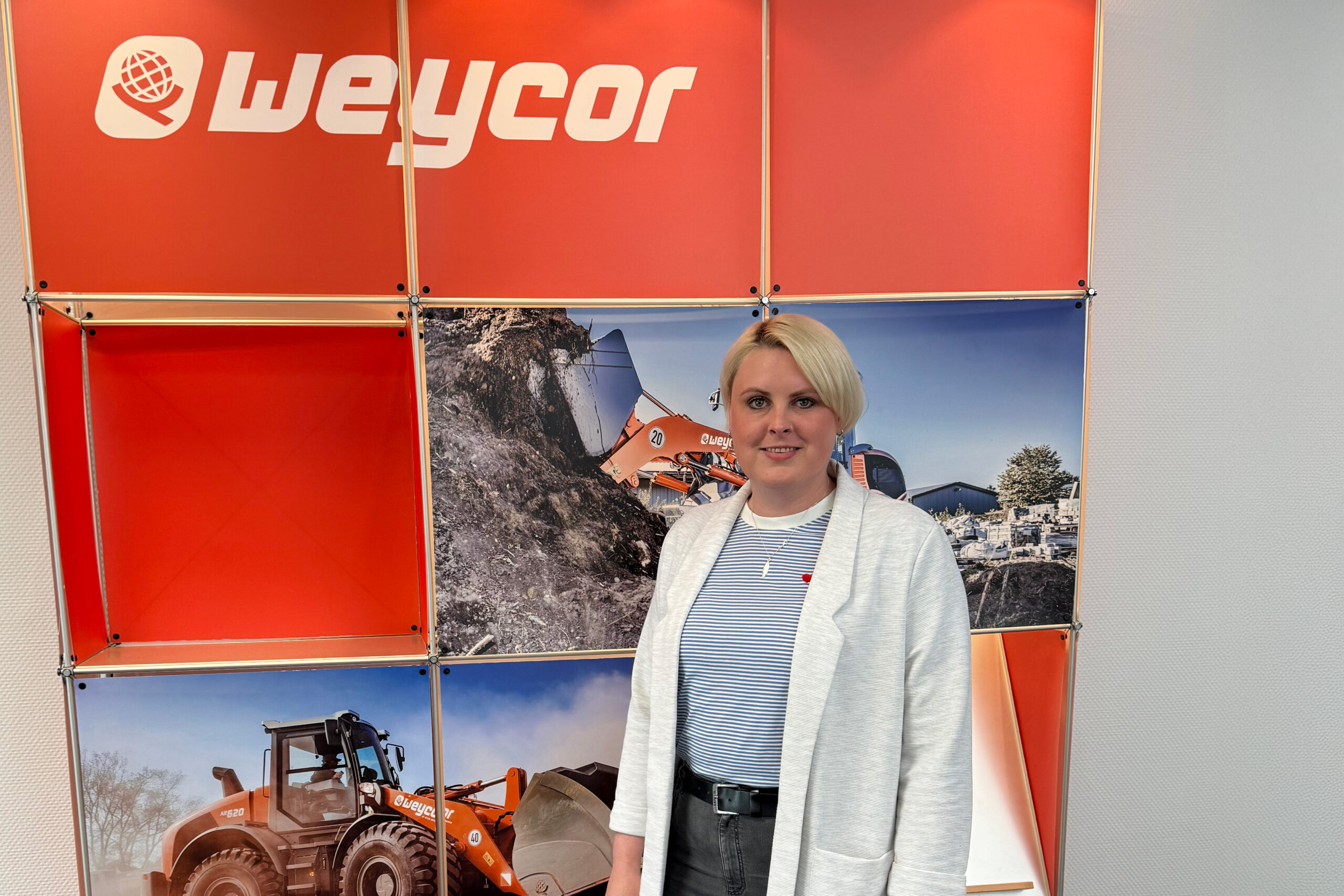 Frau mit blonden Haaren, gestreiftem Shirt und hellem Blazer steht vor einer orangen Messewand mit Weycor-Logo. Im Hintergrund Bilder von Weycor-Radladern auf Baustellen.