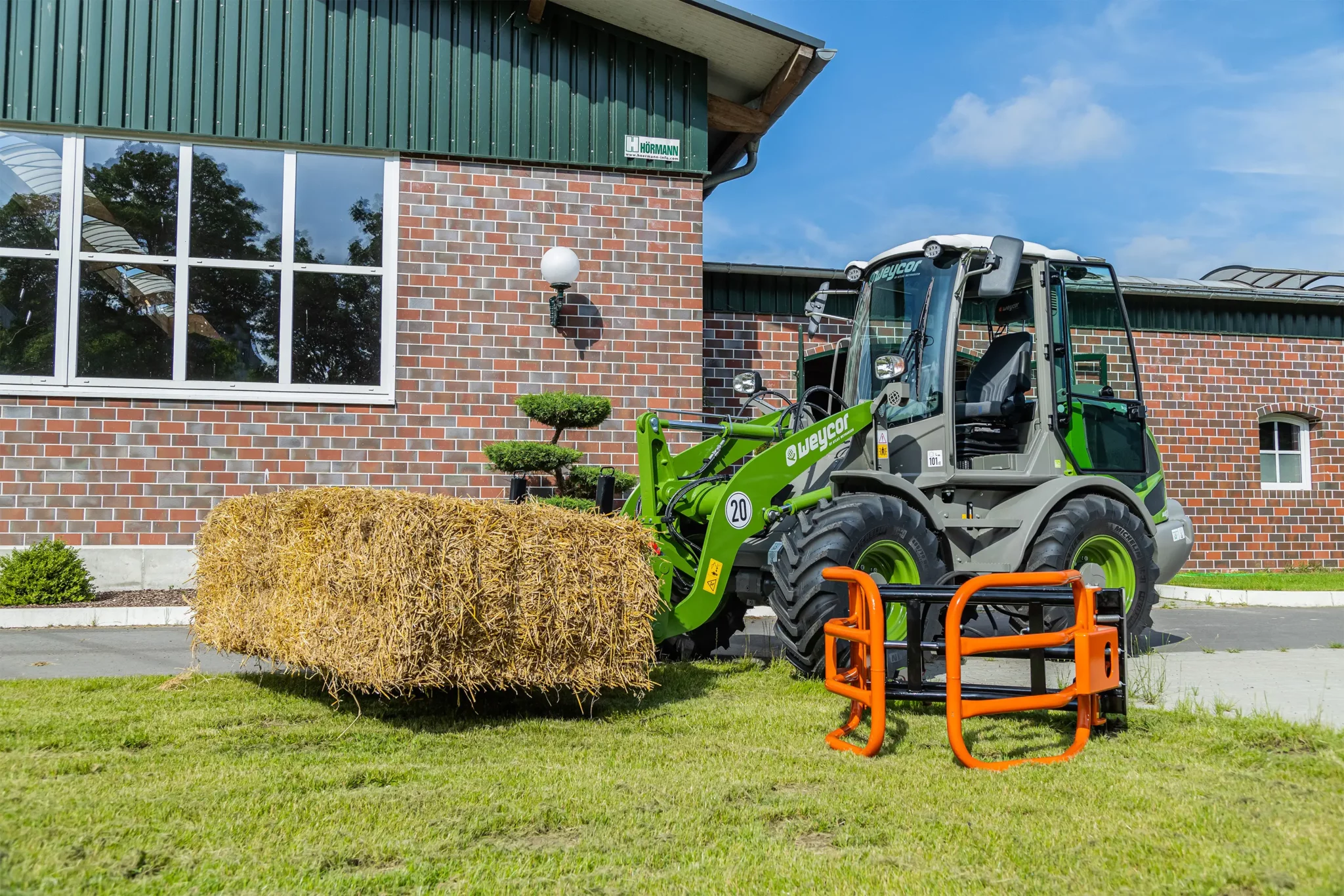 Grüner Weycor-Radlader transportiert einen großen rechteckigen Heuballen vor einem Backsteingebäude.