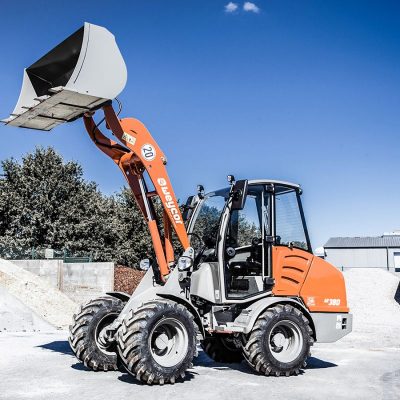 Weycor Radlader AR 380 mit hochgekipptem Frontlader, stehend auf einem Industriegelände mit großen Schüttguthaufen und Lagerhallen im Hintergrund.