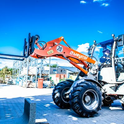 Weycor Radlader AR 380 mit Gabelaufsatz beim Materialumschlag auf einem Lagerplatz, seitliche Ansicht unter blauem Himmel.