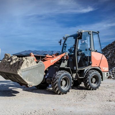 Weycor Radlader AR 320 auf Baustelle beim Transport von Sand, vor einem Haufen Bauschutt, Betonblöcken und Solarpaneelen im Hintergrund.