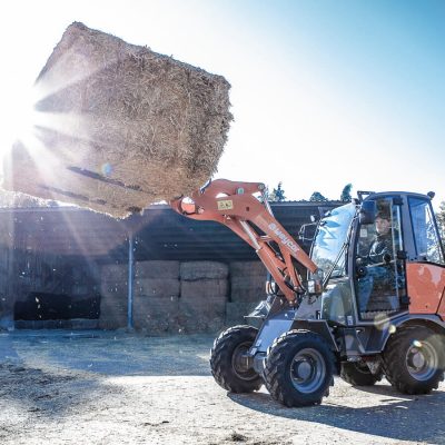 Weycor Radlader AR 320 beim Verladen eines großen Heuballens mit Palettengabel in der Landwirtschaft, Sonnenlicht im Hintergrund.