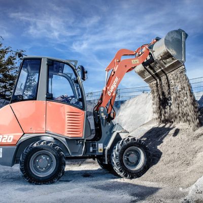 Weycor Radlader AR 320 beim Entleeren der Schaufel mit Kies oder Schotter vor einem Lagerplatz mit hohen Materialbergen.