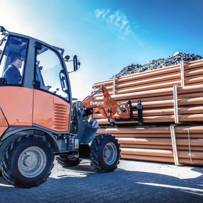 Weycor Radlader AR 320 mit Gabelaufnahme transportiert ein Bündel Kunststoffrohre auf einem Lagerplatz mit Schotterhaufen im Hintergrund.