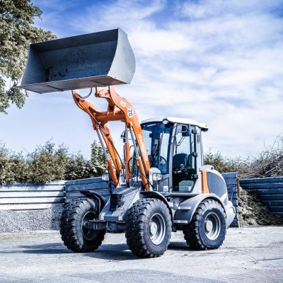 Radlader von Weycor mit hochgehobener Schaufel auf befestigtem Hof, Hintergrund mit Metallzäunen, Ästen und Büschen, blauer Himmel mit leichten Wolken.