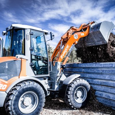 Radlader Weycor AR 420 beim Entladen von Erde oder Kompost über eine Metallbarriere, blauer Himmel mit leichten Wolken, Hintergrund mit Büschen und Erdhügeln.