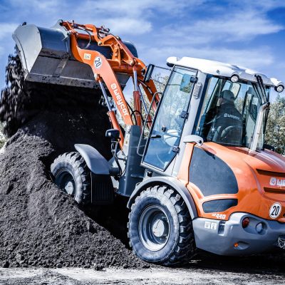 Weycor Radlader AR 440 kippt schwarze Erde auf großen Haufen, Seitenansicht mit Fahrer in der Kabine, blauer Himmel und Bäume im Hintergrund.