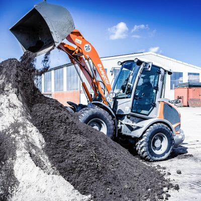 Weycor Radlader AR 440 schüttet Erde auf einen hohen Erdhaufen, Fahrer in Kabine sichtbar, im Hintergrund Industriegebäude mit Container und blauer Himmel.