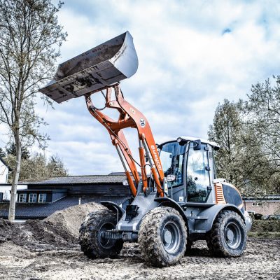 Weycor Radlader mit gehobener Schaufel auf einer schlammigen Baustelle, im Hintergrund Gebäude, Bäume und bewölkter Himmel.