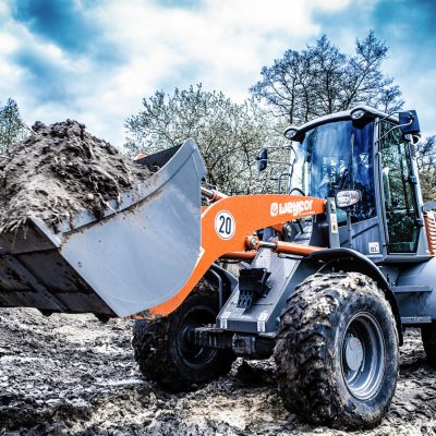 Weycor Radlader mit gefüllter Schaufel auf schlammiger Baustelle, Frontansicht mit starkem Himmel, Bäume im Hintergrund.