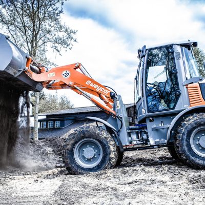 Weycor Radlader schüttet Erdmaterial aus erhobener Schaufel auf matschigem Baustellenboden, Seitenansicht, Bäume und Gebäude im Hintergrund.