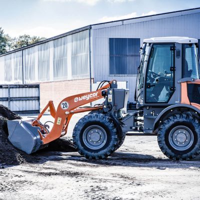 Weycor Radlader AR 480S im Einsatz beim Aufnehmen von Erde vor einer Industriehalle, Seitenansicht.