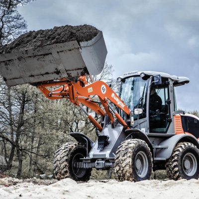 Weycor Radlader AR 520 beim Heben einer vollen Schaufel mit Erde, Einsatz auf sandigem Boden in Waldnähe, bewölkter Himmel.