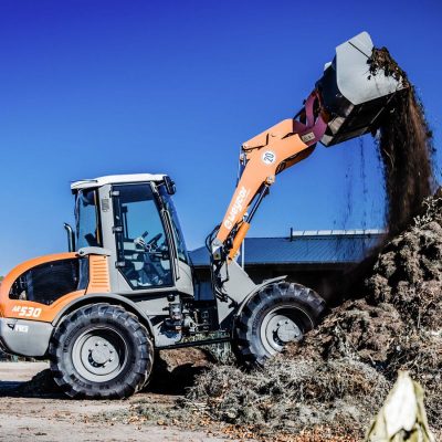 Weycor Radlader AR 530 beim Entladen von organischem Material auf einen Komposthaufen, Seitenansicht von links, sonniges Wetter mit blauem Himmel.