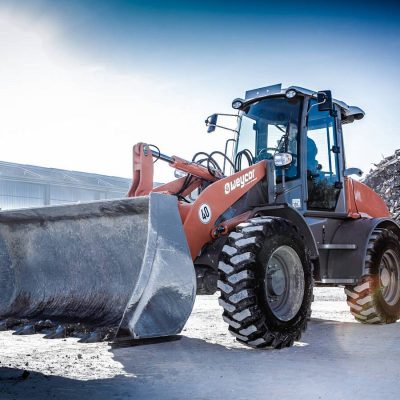 Weycor Radlader AR 560 mit großer Schaufel auf Baustelle, Aufnahme von schräg vorne links, im Hintergrund Schutthaufen und Lagerhallen, bei hellem Tageslicht.