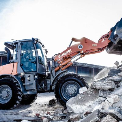 Weycor Radlader AR 560 mit hochgefahrener Schaufel arbeitet auf einem Bauschutthaufen aus Beton- und Ziegelbruch, Fahrer sichtbar in der Kabine, im Hintergrund Industriehallen und freier Himmel.