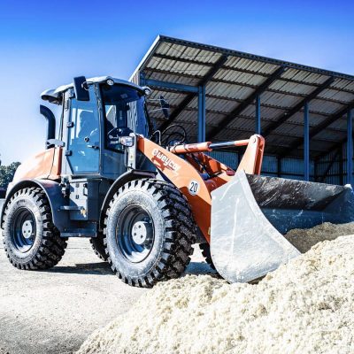 Weycor wheel loader with front shovel, taken from the side, shovel immersed in a pile of sand or bulk material, in the background an open metal warehouse, clear blue sky.