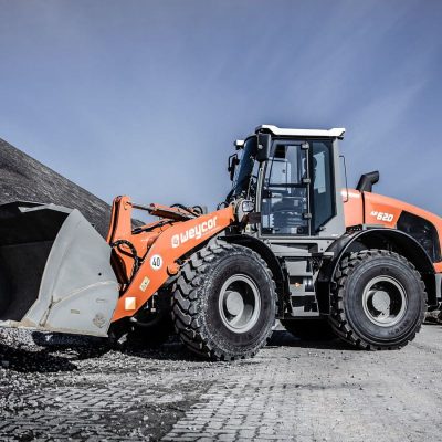 Weycor Radlader AR 640 auf Schotterplatz, seitlich fotografiert mit angehobener Schaufel, Schaufelzähne sichtbar, orange-graues Fahrzeug, im Hintergrund aufgetürmte große Steine, klarer blauer Himmel.