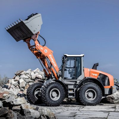 Weycor Radlader AR 640 auf Schotterplatz, seitlich fotografiert mit angehobener Schaufel, Schaufelzähne sichtbar, orange-graues Fahrzeug, im Hintergrund aufgetürmte große Steine, klarer blauer Himmel.