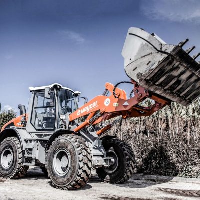 Weycor Radlader AR 680 auf Baustelle, seitlich aufgenommen mit angehobener Schaufel, grobe Zähne an der Schaufel, orange-graue Maschine, große Profilreifen, Hecke und blauer Himmel im Hintergrund.