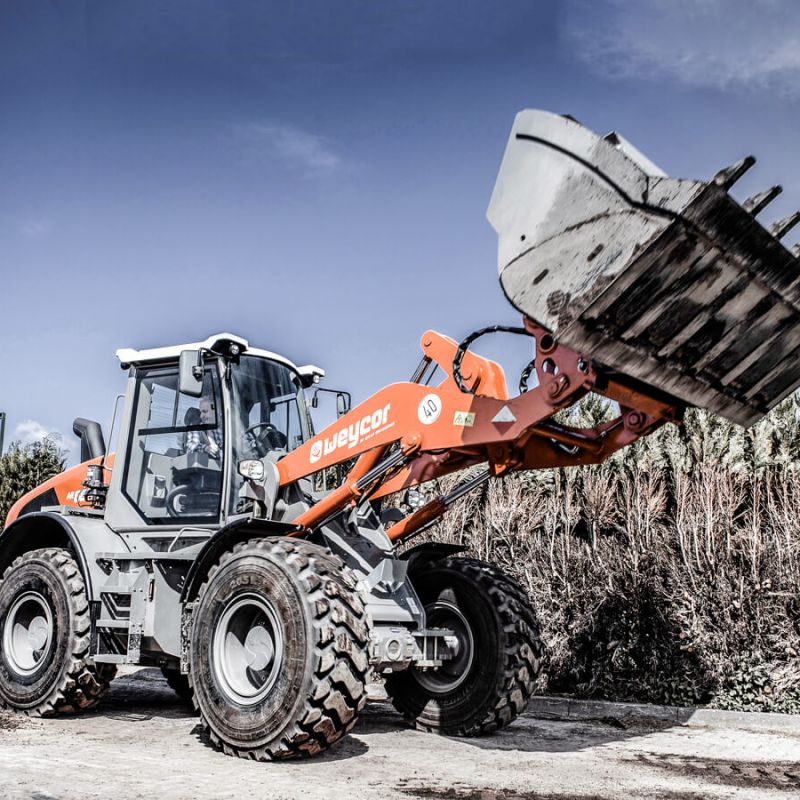 Ładowarka kołowa Weycor AR 680 na placu budowy, sfotografowana z boku z podniesioną łyżką, grube zęby na łyżce, pomarańczowo-szara maszyna, duże opony profilowane, żywopłot i błękitne niebo w tle.